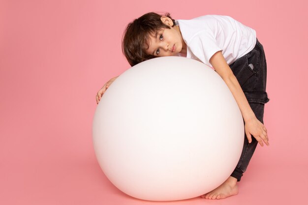 Una vista frontal chico lindo en camiseta blanca jugando con pelota redonda blanca en el piso rosa
