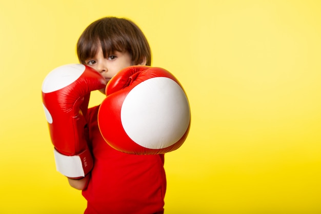 Una vista frontal chico lindo en camiseta roja y con guantes de boxeo rojos en la pared amarilla