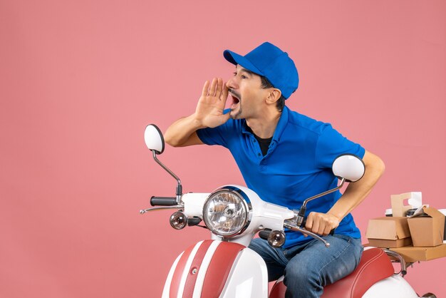 Vista frontal del chico de mensajería confundido con sombrero sentado en scooter y llamando a alguien sobre fondo melocotón pastel