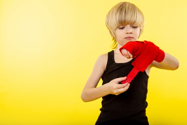 Una vista frontal chico rubio en camiseta negra y tejido rojo alrededor de su mano en la pared amarilla