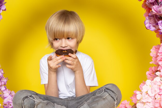 Una vista frontal chico rubio comiendo donas en camiseta blanca en el piso amarillo