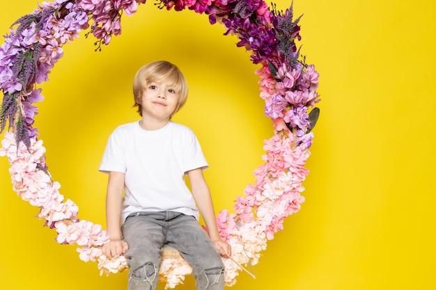 Una vista frontal chico rubio dulce lindo adorable en camiseta blanca sentada en la flor hecha soporte en el escritorio amarillo