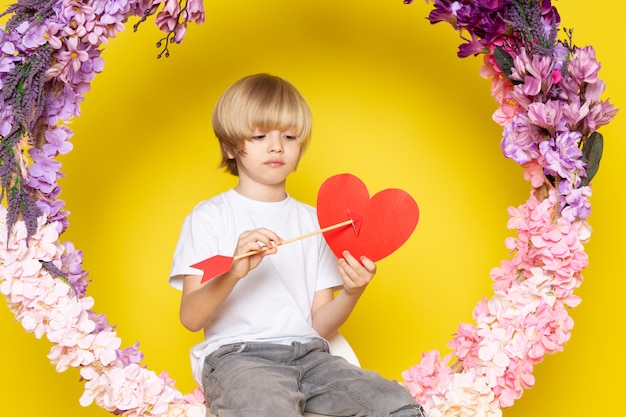 Una vista frontal chico rubio lindo adorable en camiseta blanca con forma de corazón en el escritorio hecho de flores en el piso amarillo