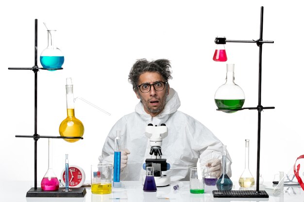 Vista frontal científico loco en traje de protección especial sentado alrededor de la mesa con soluciones en la enfermedad de escritorio blanco virus de la ciencia del laboratorio covid
