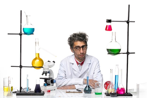 Vista frontal científico masculino en traje médico blanco sentado frente a la mesa con soluciones