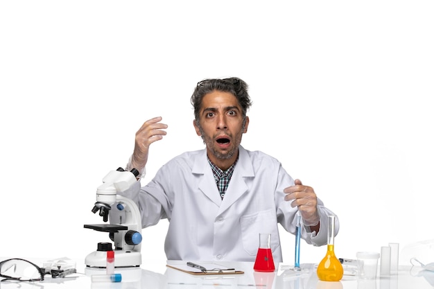 Vista frontal científico de mediana edad en traje médico sentado alrededor de la mesa con soluciones