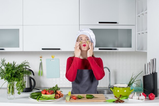 Vista frontal de la cocinera con sombrero de cocinero poniendo rodajas de pepino en la cara en la cocina