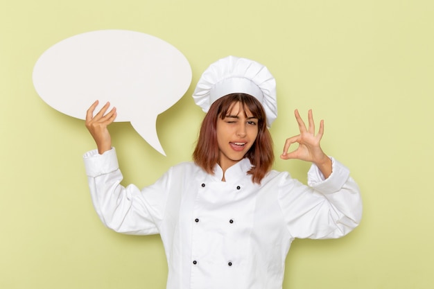 Vista frontal cocinera en traje de cocinero blanco con un gran cartel blanco en el escritorio verde