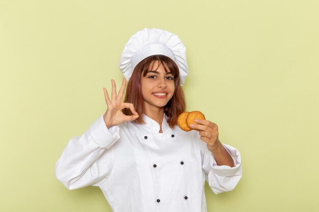 Vista frontal cocinera en traje de cocinero blanco sosteniendo galletas de azúcar en el escritorio verde