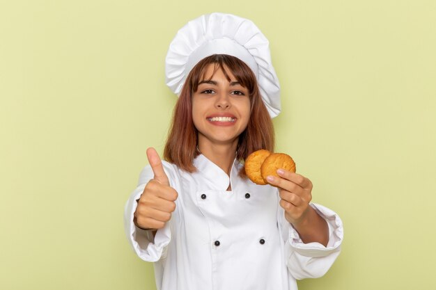 Vista frontal cocinera en traje de cocinero blanco sosteniendo galletas de azúcar sobre una superficie verde