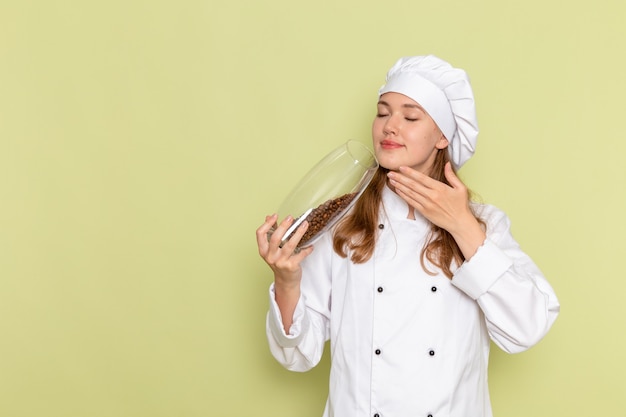 Vista frontal de la cocinera en traje de cocinero blanco sosteniendo lata con semillas de café marrón en la pared verde