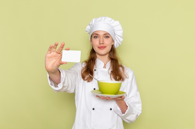 Vista frontal de la cocinera en traje de cocinero blanco sosteniendo la placa y la tarjeta en la pared verde