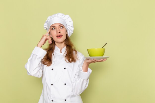 Vista frontal de la cocinera en traje de cocinero blanco sosteniendo placa verde con plato en la pared verde