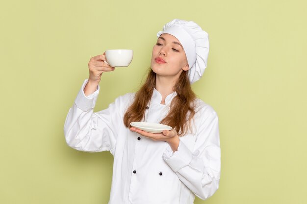 Vista frontal de la cocinera en traje de cocinero blanco sosteniendo una taza de café en la pared verde