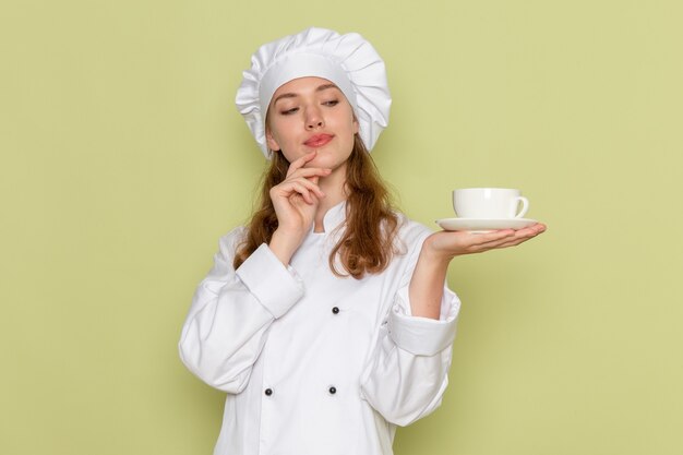 Vista frontal de la cocinera en traje de cocinero blanco sosteniendo una taza de café en la pared verde