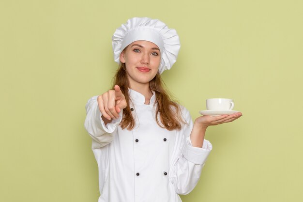 Vista frontal de la cocinera en traje de cocinero blanco sosteniendo una taza de café y sonriendo en la pared verde