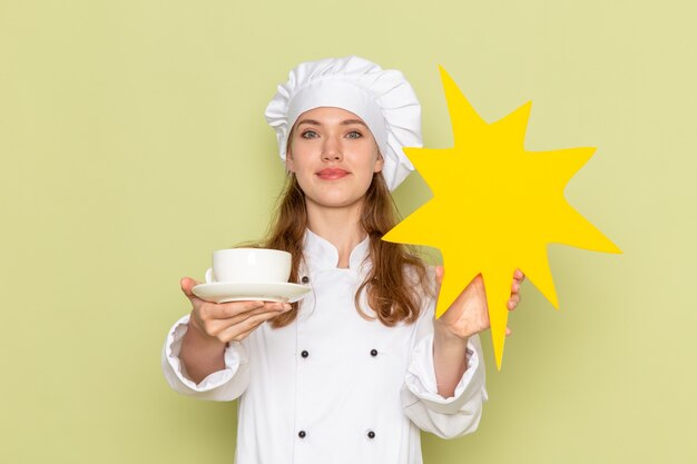 Vista frontal de la cocinera en traje de cocinero blanco sosteniendo la taza y el cartel amarillo en la pared verde