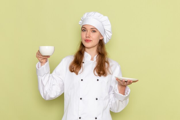 Vista frontal de la cocinera en traje de cocinero blanco sosteniendo la taza y el plato en la pared verde