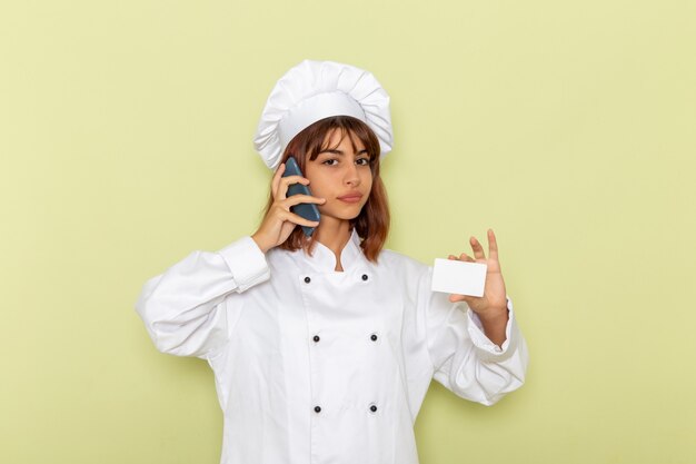 Vista frontal cocinera en traje de cocinero blanco con tarjeta hablando por teléfono en superficie verde