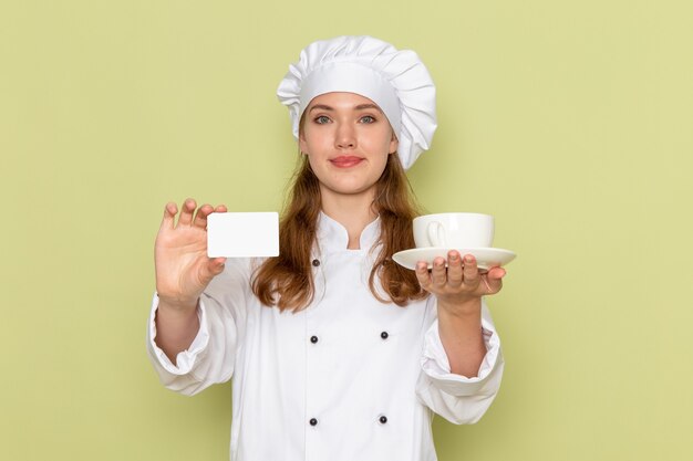 Vista frontal de la cocinera en traje de cocinero blanco con taza y tarjeta en la pared verde