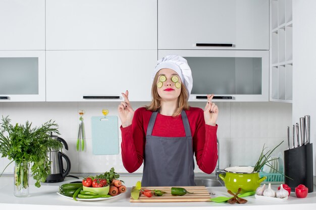 Vista frontal de la cocinera en uniforme poniendo rodajas de pepino en la cara haciendo un signo de buena suerte