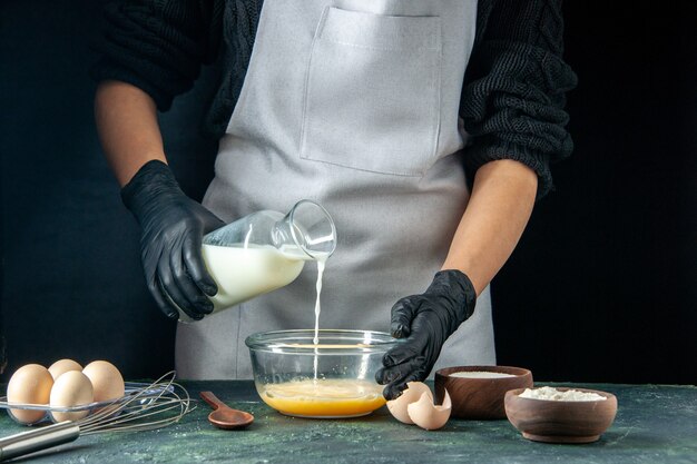 Vista frontal cocinera vertiendo leche en los huevos en pasteles oscuros pastel pastel trabajador cocina masa trabajo hotcakes