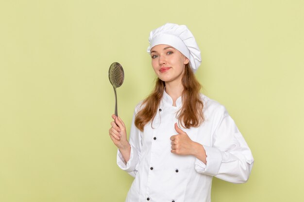 Vista frontal de la cocinera vistiendo traje de cocinero blanco sosteniendo una cuchara de plata grande en la pared verde
