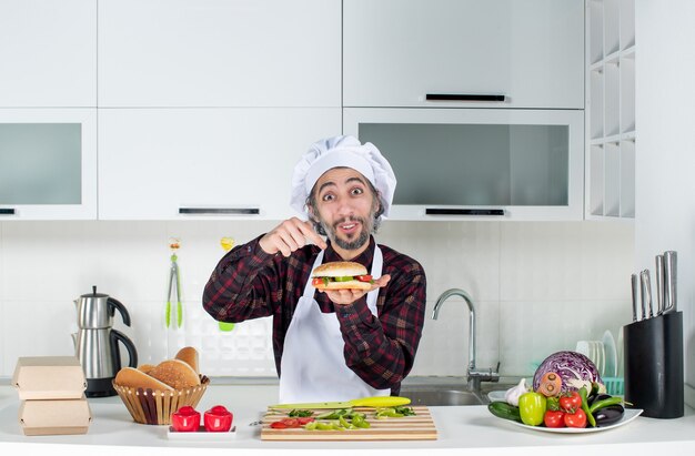 Vista frontal del cocinero macho sosteniendo una gran hamburguesa de pie detrás de la mesa de la cocina