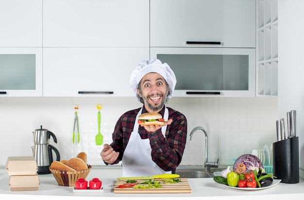 Vista frontal del cocinero macho sosteniendo una sabrosa hamburguesa de pie detrás de la mesa de la cocina