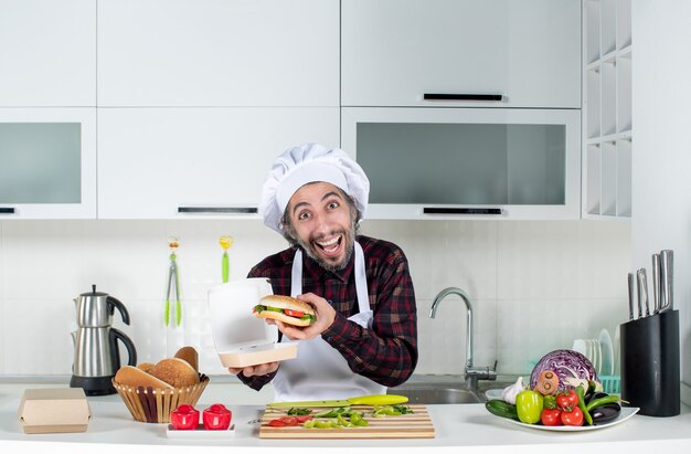 Vista frontal del cocinero macho tomando una gran hamburguesa de la caja de pie detrás de la mesa de la cocina