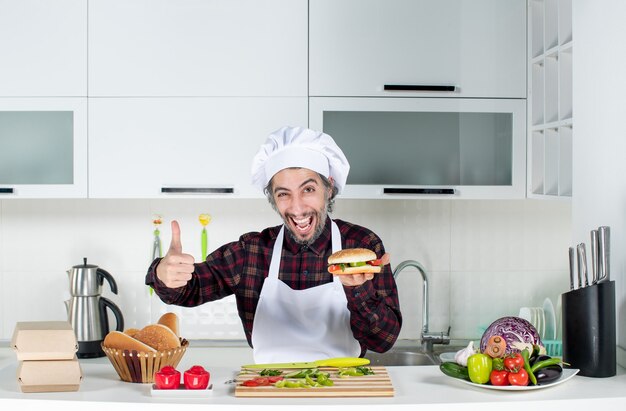 Vista frontal del cocinero masculino dando pulgares arriba sosteniendo una hamburguesa de pie detrás de la mesa de la cocina