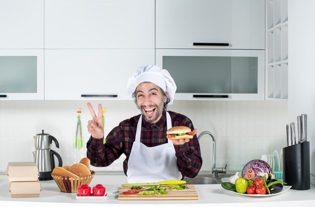 Vista frontal del cocinero masculino haciendo el signo de la victoria sosteniendo una hamburguesa de pie detrás de la mesa de la cocina