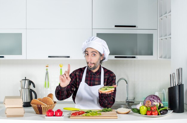 Vista frontal del cocinero masculino preguntado haciendo hamburguesas de pie detrás de la mesa de la cocina