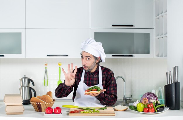 Vista frontal del cocinero masculino sosteniendo una deliciosa hamburguesa de pie detrás de la mesa de la cocina