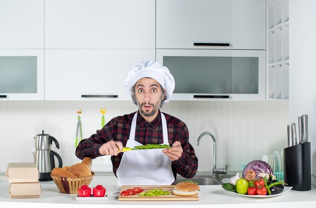 Vista frontal del cocinero con los ojos abiertos sosteniendo un cuchillo cortando verduras en la cocina