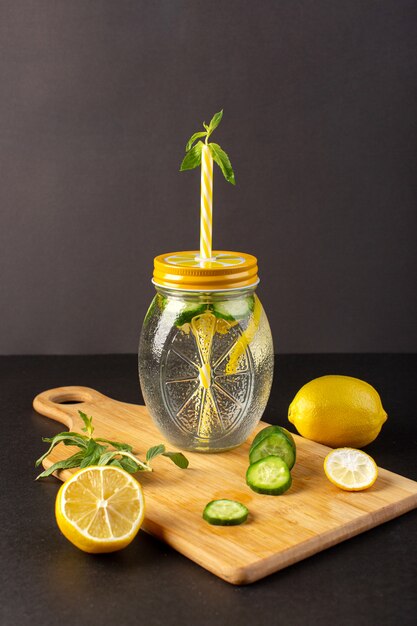 Una vista frontal cóctel de limón bebida fresca fresca dentro de la taza de vidrio en rodajas y limones enteros pepinos junto con flores de paja en el fondo oscuro cóctel bebida fruta