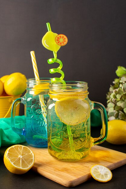 Una vista frontal cóctel de limón bebida fresca fresca dentro de vasos de vidrio en rodajas y limones enteros pajitas de colores sobre el fondo oscuro cóctel bebida fruta