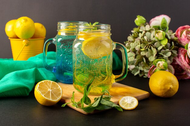 Una vista frontal cóctel de limón refresco fresco dentro de vasos de vidrio en rodajas y limones enteros junto con flores en el fondo oscuro cóctel bebida fruta