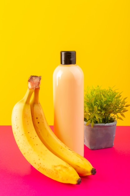 Una vista frontal de color crema botella de champú de plástico puede con tapa negra aislada con plátanos y pequeña planta en el fondo rosa-amarillo cosméticos belleza cabello