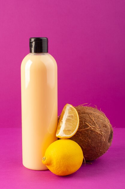 Una vista frontal de color crema botella de champú de plástico puede con tapa negra junto con limones y coco aislado en el fondo púrpura cosméticos belleza cabello