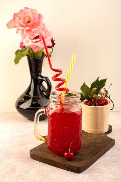 Una vista frontal de color rojo cereza cóctel con pajitas dentro de una pequeña lata de enfriamiento fresco en un escritorio de madera junto con cerezas frescas y flores y un escritorio rosa