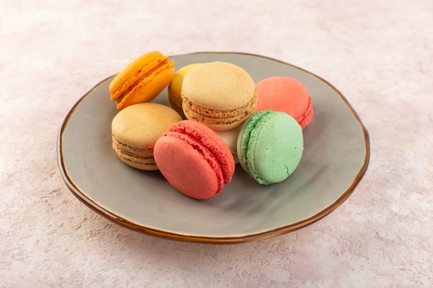 Una vista frontal de coloridos macarons franceses dentro de la placa en el escritorio rosa pastel dulce de azúcar de galletas