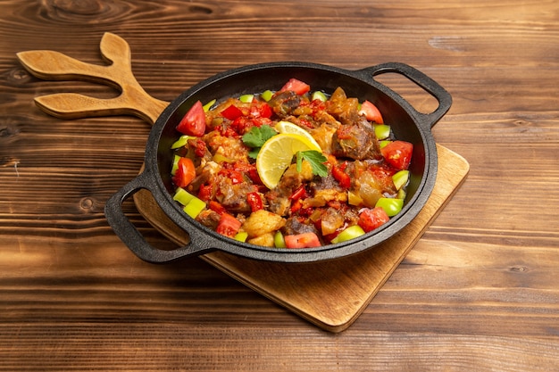 Vista frontal comida vegetal cocida con carne y pimientos en rodajas dentro de la sartén sobre superficie de madera marrón