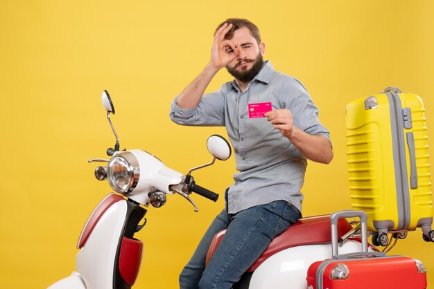 Vista frontal del concepto de viaje con hombre barbudo confiado joven sentado en la motocicleta mostrando tarjeta bancaria haciendo gesto de anteojos en amarillo