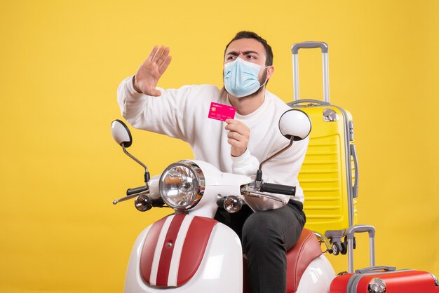 Vista frontal del concepto de viaje con un joven nervioso con máscara médica sentado en una motocicleta con una maleta amarilla y sosteniendo una tarjeta bancaria