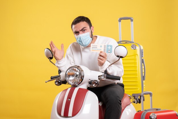 Vista frontal del concepto de viaje con un joven pensador en máscara médica sentado en una motocicleta con una maleta amarilla y sosteniendo un boleto