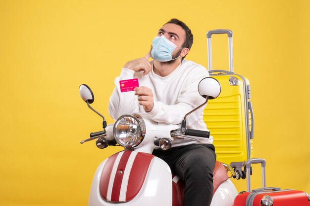 Vista frontal del concepto de viaje con un joven pensador en máscara médica sentado en una motocicleta con una maleta amarilla y sosteniendo una tarjeta bancaria