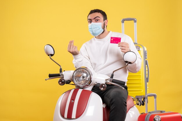 Vista frontal del concepto de viaje con un joven sorprendido con máscara médica sentado en una motocicleta con una maleta amarilla y sosteniendo una tarjeta bancaria