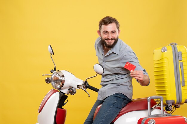 Vista frontal del concepto de viaje con sonriente joven sentado en motocicleta con maletas con tarjeta bancaria en amarillo