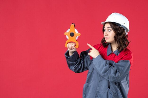 Vista frontal de la confiada arquitecta en uniforme con casco sosteniendo cinta métrica y apuntando a la pared roja aislada